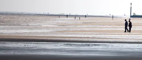 Sun sea sand shadow 2 This landscape photograph depicts Sutton on Sea Beach in Lincolnshire, England, on a late spring morning. The main subject, titled "Sun sea sand shadow 2," shows people walking along the expansive sandy beach as the sunlight creates shadow patterns across the sand and shallow tidal pools. The gentle waves of the North Sea can be seen in the background, while distant figures stroll along the shore, illustrating the quiet coastal charm of this United Kingdom location. A prominent beach marker is visible near the horizon, adding a distinctive feature to the seafront scene and enhancing the local identity of Sutton on Sea.